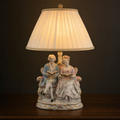 Lit porcelain figurine table lamp with two figures reading together on a settee, ivory pleated shade