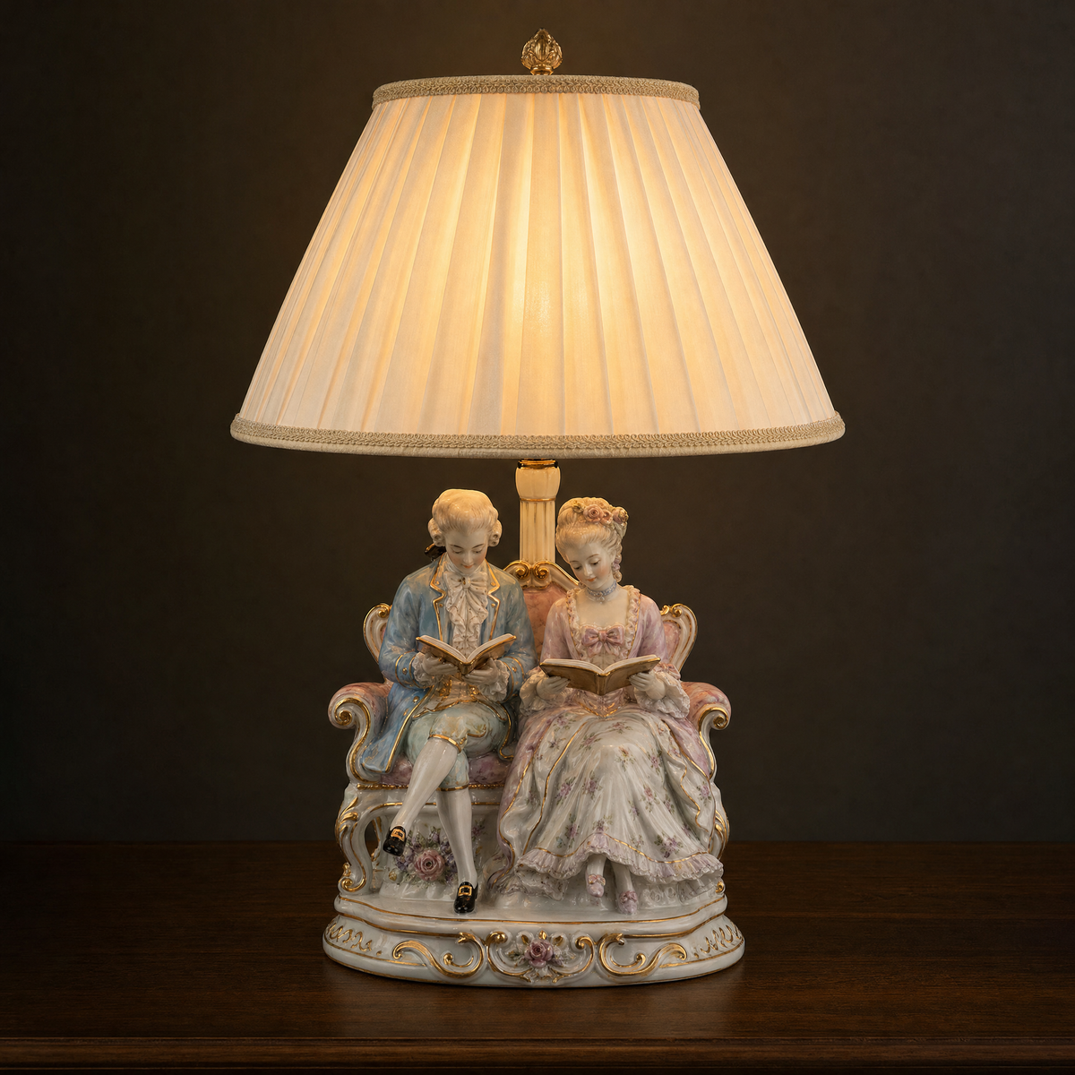 Lit porcelain figurine table lamp with two figures reading together on a settee, ivory pleated shade