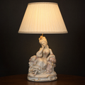 Lit porcelain figurine table lamp with a single seated woman in a court gown resting against a cushion